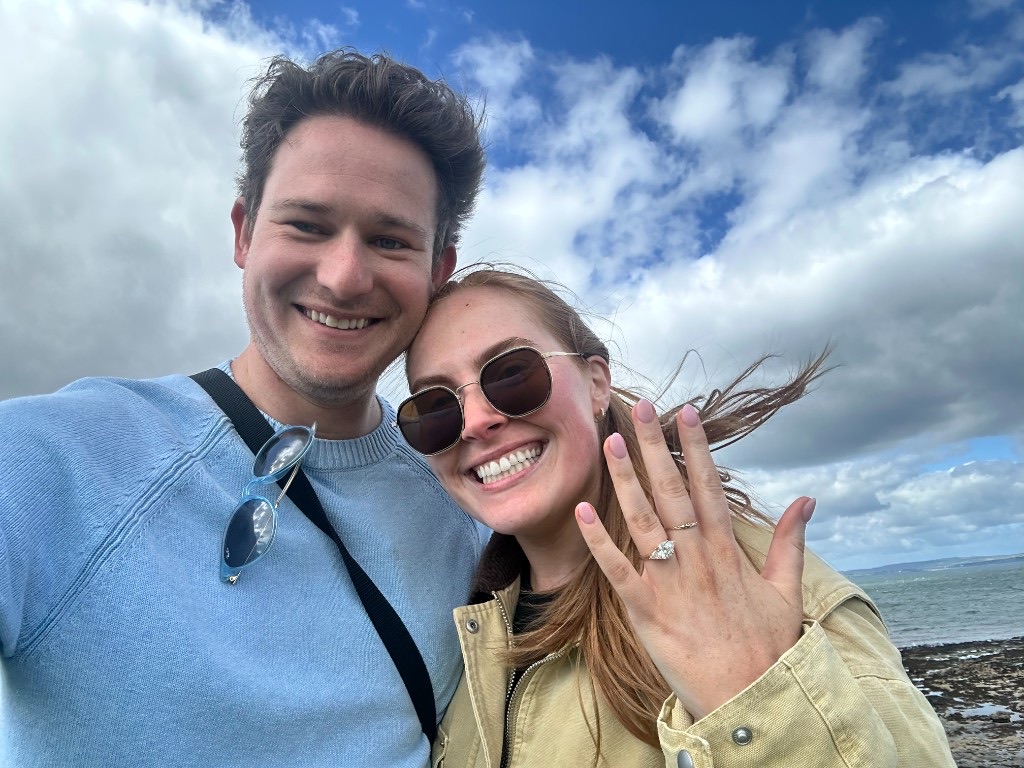 Laura and Andrew smiling, showing Laura's engagement ring by the coast